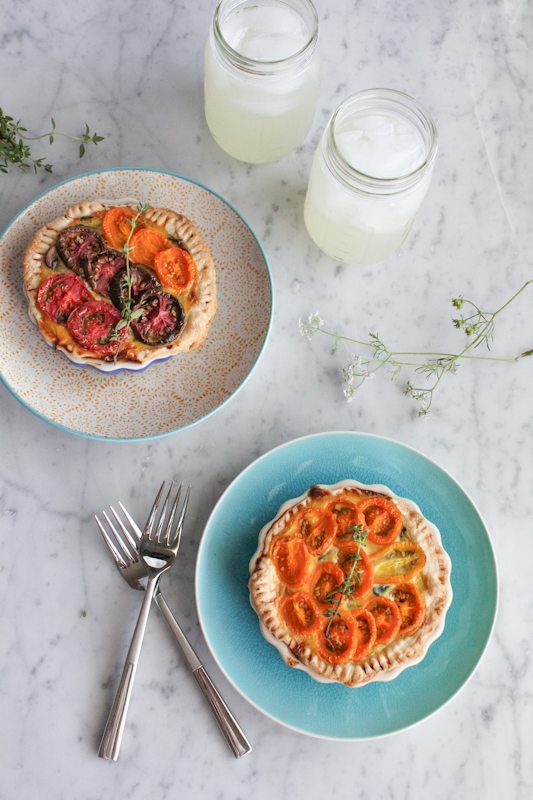 Two plates of Heirloom tomato tarts, two forks, and two glass jars with beverages.
