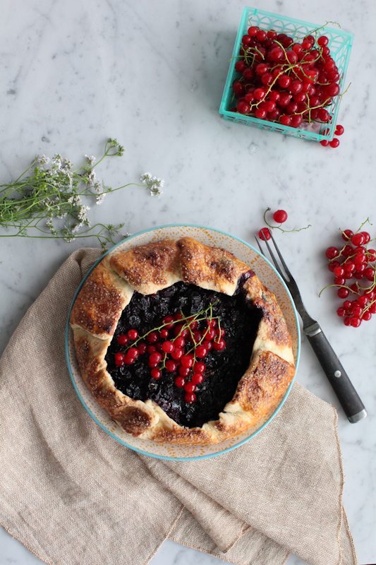 A mini berry galette on a plate with a basket of berries, and flowers.