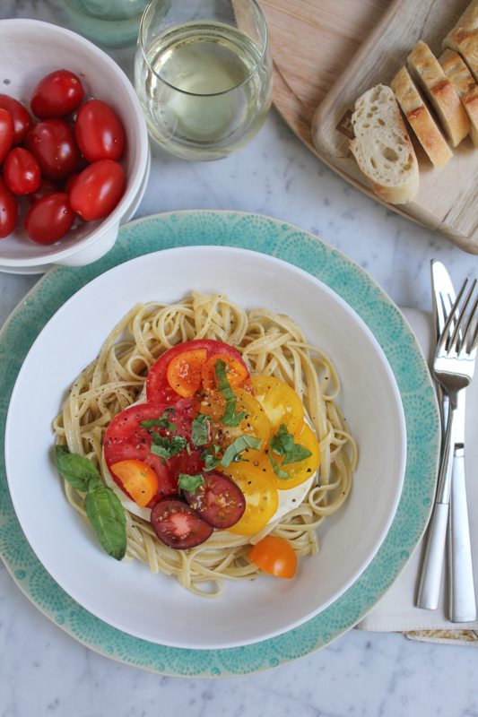 Summer Caprese pasta salad in a bowl with a small bowl of tomatoes, a glass of white wine, a wooden board with sliced bread, and silverware.