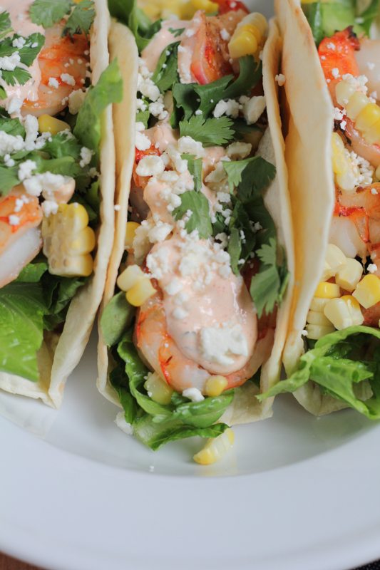 Grilled Shrimp Tacos with Mexican Crema topped with grilled corn, cilantro, and cheese, on a white plate.