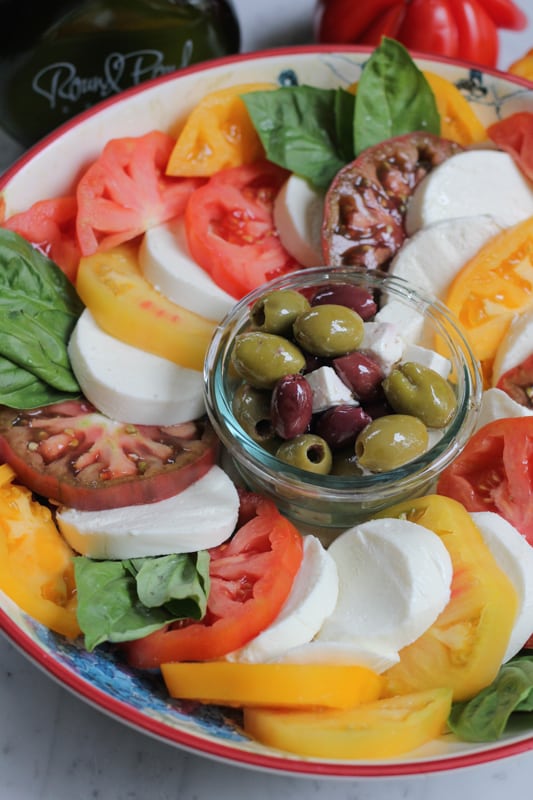 Photo of a platter of Simple Caprese Salad with a bowl of olives and feta in the center.