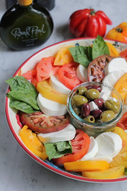 A platter of Simple Caprese Salad with a bowl of olives and feta, and a bottle of olive oil and tomatos in the background.
