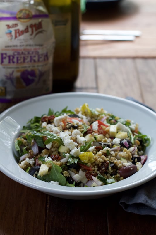 A bowl of Mediterranean Freekeh Salad with a bag of freekeh grains and olive oil in the background.