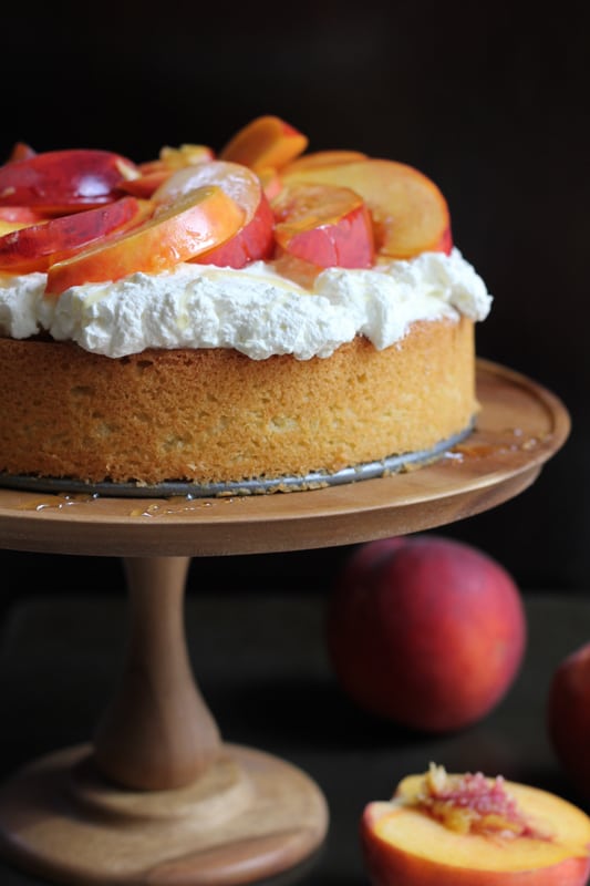 Olive Oil Honey Cake With Fresh Peaches on a wooden cake stand, surrounded by peaches.