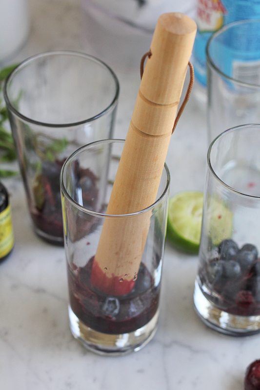 Blueberries being muddled at the bottom of a glass for blueberry cherry mojitos.