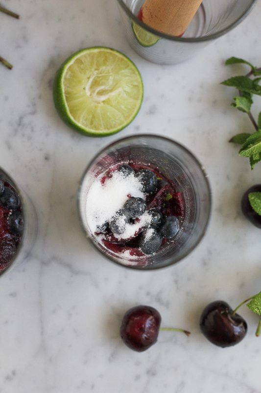 Blueberries and sugar at the bottom of a glass with half a lime, mint sprigs, and cherries.