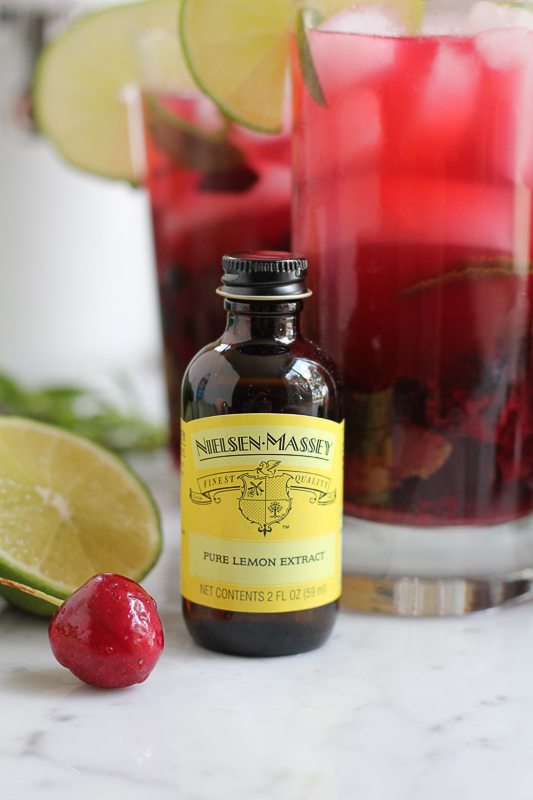 A bottle of lemon extract with glasses of blueberry cherry mojitos in the background.