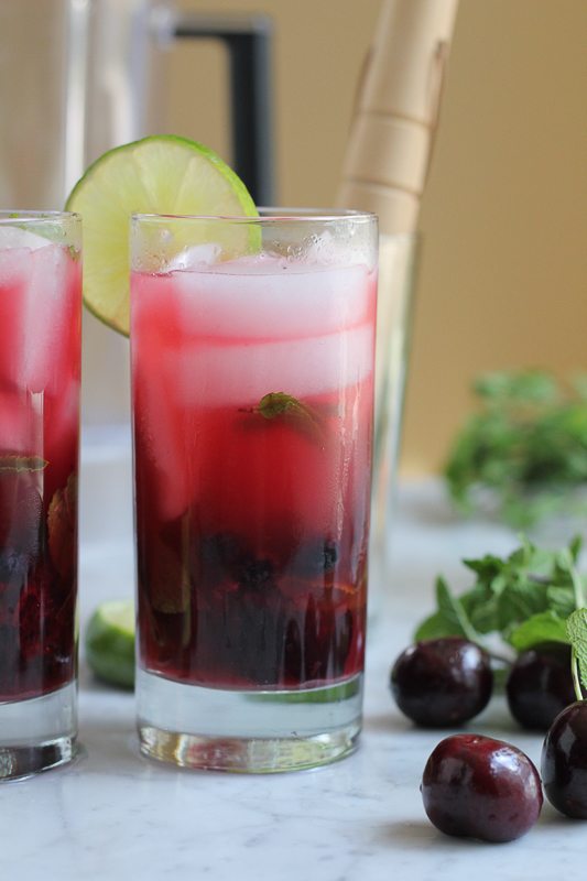 A blueberry cherry mojito in a glass, garnished with a slice of lime, with cherries and mint.