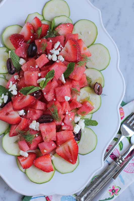 Zucchini watermelon and feta salad on a white plate with a napkin and silverware.