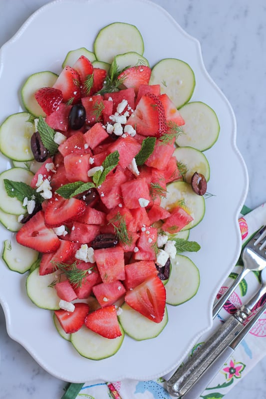 Zucchini watermelon and feta salad on a white plate with napkins and silverware.