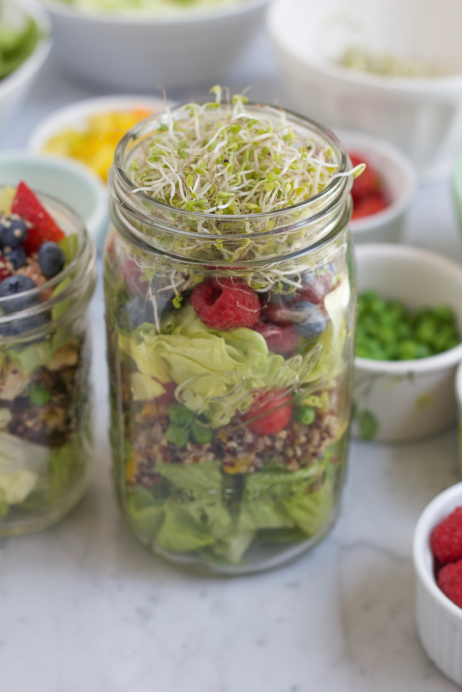 Two open jars of Mason Jar Salad with bowls of ingredients in the background.