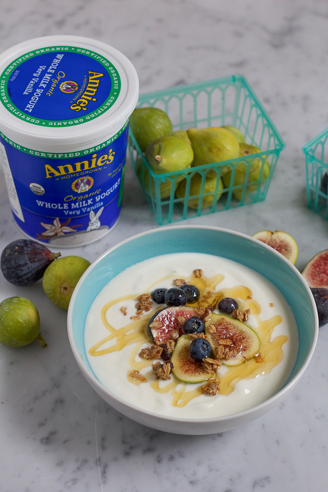 A bowl of yogurt topped with fruit, granola, and honey, with yogurt container and figs.
