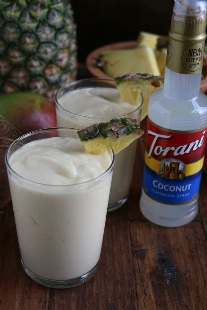 Two glasses of Refreshing Tropical Coconut Smoothie with a pineapple, mangos, and bottle of coconut syrup.