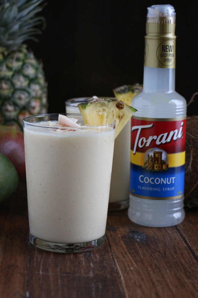 Two glasses of Refreshing Tropical Coconut Smoothie topped with coconut flakes, with a pineapple, mangos and a bottle of coconut syrup.