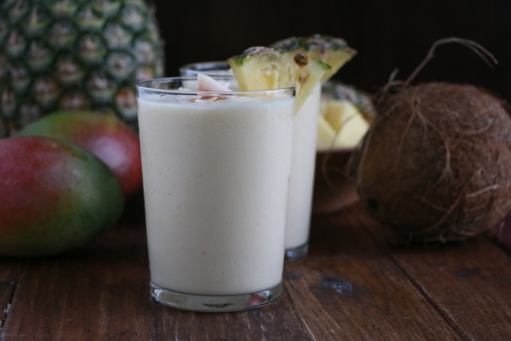 Two glasses of Refreshing Tropical Coconut Smoothie, with a pineapple, coconut, and mangos in the background.
