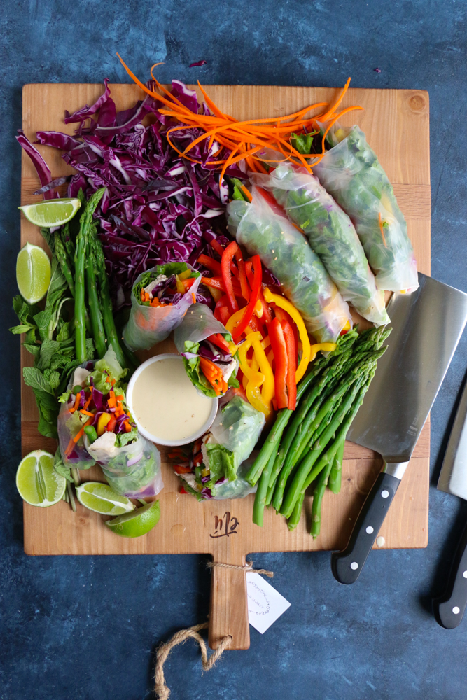 A board of chopped vegetables, Chicken and Veggie Spring Rolls, a bowl of dipping sauce, and a knife.