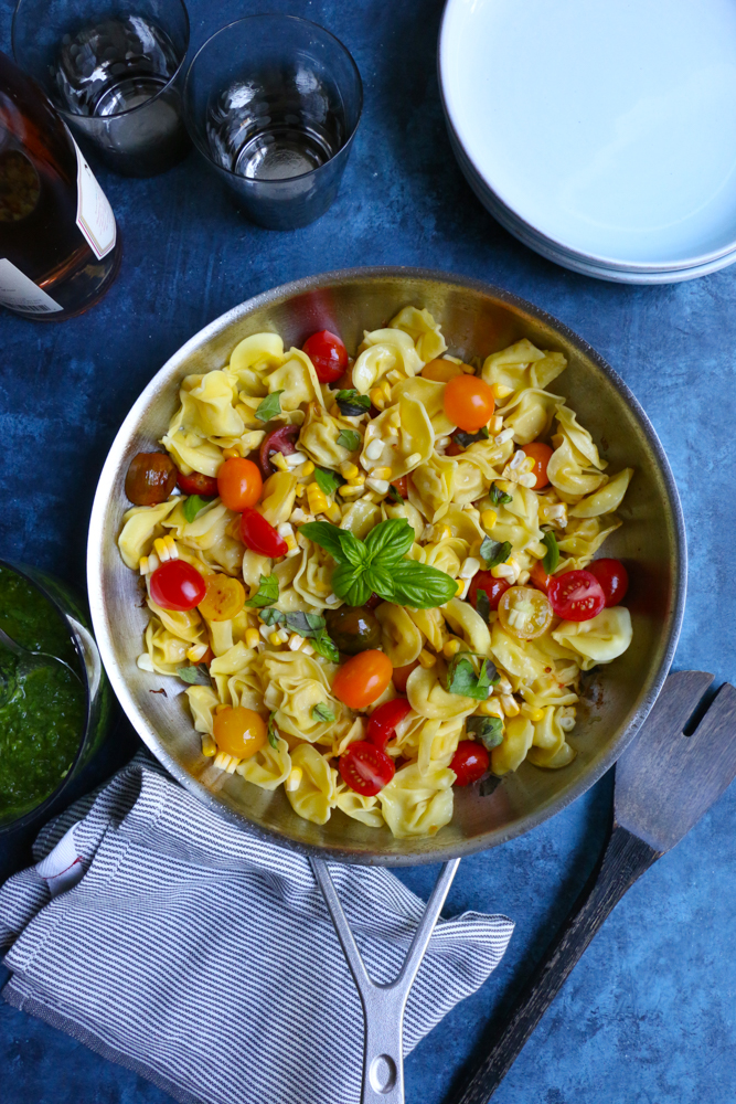 A skillet of Summer Tortellini Pasta Salad with a bottle of wine, glasses, bowls, a napkin, and a serving spoon.
