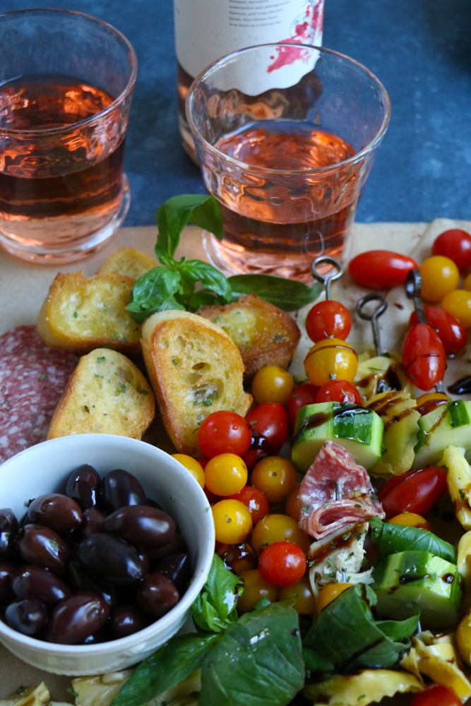 Summer Tortellini Pasta Salad on skewers, a bowl of olives, slices of bread, and two glasses of wine.